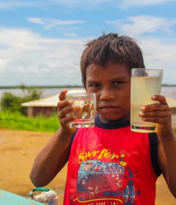 A child looking at the camera during the installation of a tank of 45,000 liters, which will provide access to potable water to an estimated 1,500 people per month, among which 500 indigenous people of the Warao ethnic group stand out, Cambalache Community, Bolívar state, on July 20, 2020. In his right hand, the child holds water drawn from the newly installed water tank; in his left, he holds water from the river, which had been the community's water source prior to the tank installation.