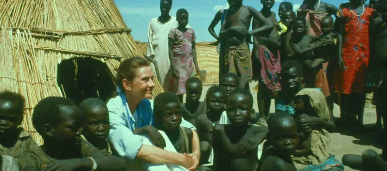 United Nations good will ambassador Audrey Hepburn sitting among scores of war affected children and women in southern Sudan, in front a hut while other are standing looking at the visitor