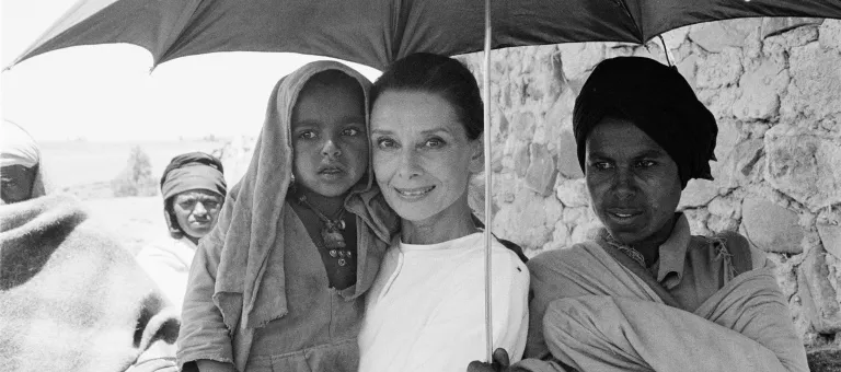 Others nearby, UNICEF Goodwill Ambassador Audrey Hepburn carries a small girl, standing under an umbrella held by an older boy, in the northern town of Mehal Meda in Shoa Province. Ms. Hepburn was in the town to visit a 'cash-for-food' project that includes a sheep-breeding component to provide wool for weaving and blanket-making.
