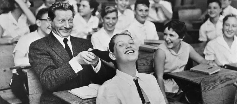 UNICEF Goodwill Ambassador Danny Kaye playfully pulls the hair of a girl seated in front of him, in a classroom filled with laughing children, during a visit to his primary-school alma mater, Public School 149 in the borough of Brooklyn in New York City. Mr. Kaye was visiting the school to help raise funds for UNICEF by encouraging children to take UNICEF coin collection boxes along on their annual 'Trick-or-Treat' rounds, door-to-door, during Halloween.