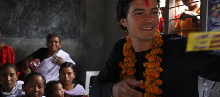 British actor Orlando Bloom visits children and women who are ethnic Tharu at the Sayapatri Community Organization in Dibyanagar Village in Chitwan District. UNICEF supported the construction of the single-room building where growth monitoring and other child support activities are held. Mr. Bloom is wearing garlands of fresh flowers around his neck and has a red 'tikka' mark on his forehead, both are local symbols of welcome.