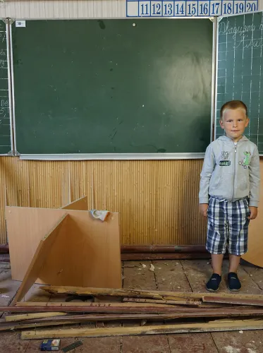 Kolya, 5 jaar, staat in een klaslokaal van de basisschool in Mostysche, Oekraïne, die zwaar beschadigd werd tijdens de beschietingen in februari en maart 2022. Op het schoolbord staat de datum van de laatste les op de school - 23 februari 2022. © UNICEF/UN0691560/Hrom