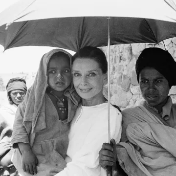 Others nearby, UNICEF Goodwill Ambassador Audrey Hepburn carries a small girl, standing under an umbrella held by an older boy, in the northern town of Mehal Meda in Shoa Province. Ms. Hepburn was in the town to visit a 'cash-for-food' project that includes a sheep-breeding component to provide wool for weaving and blanket-making.